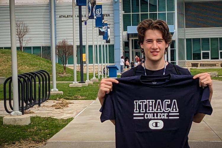 Caleb Ghia stands in front of the Ithaca College Athletic Center, holding up an Ithaca College T-shirt and smiling
