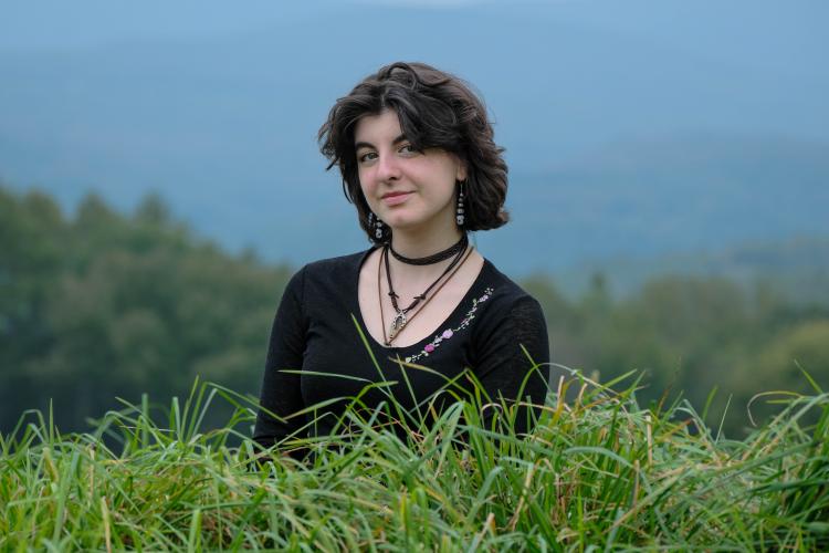 Photograph of Navia Garcia, a young person in a black shirt with necklaces, standing amongst tall grasses.