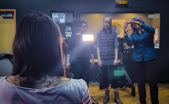 a group of people stand by a camera and lights while a person sits in a chair, in a lighting workshop