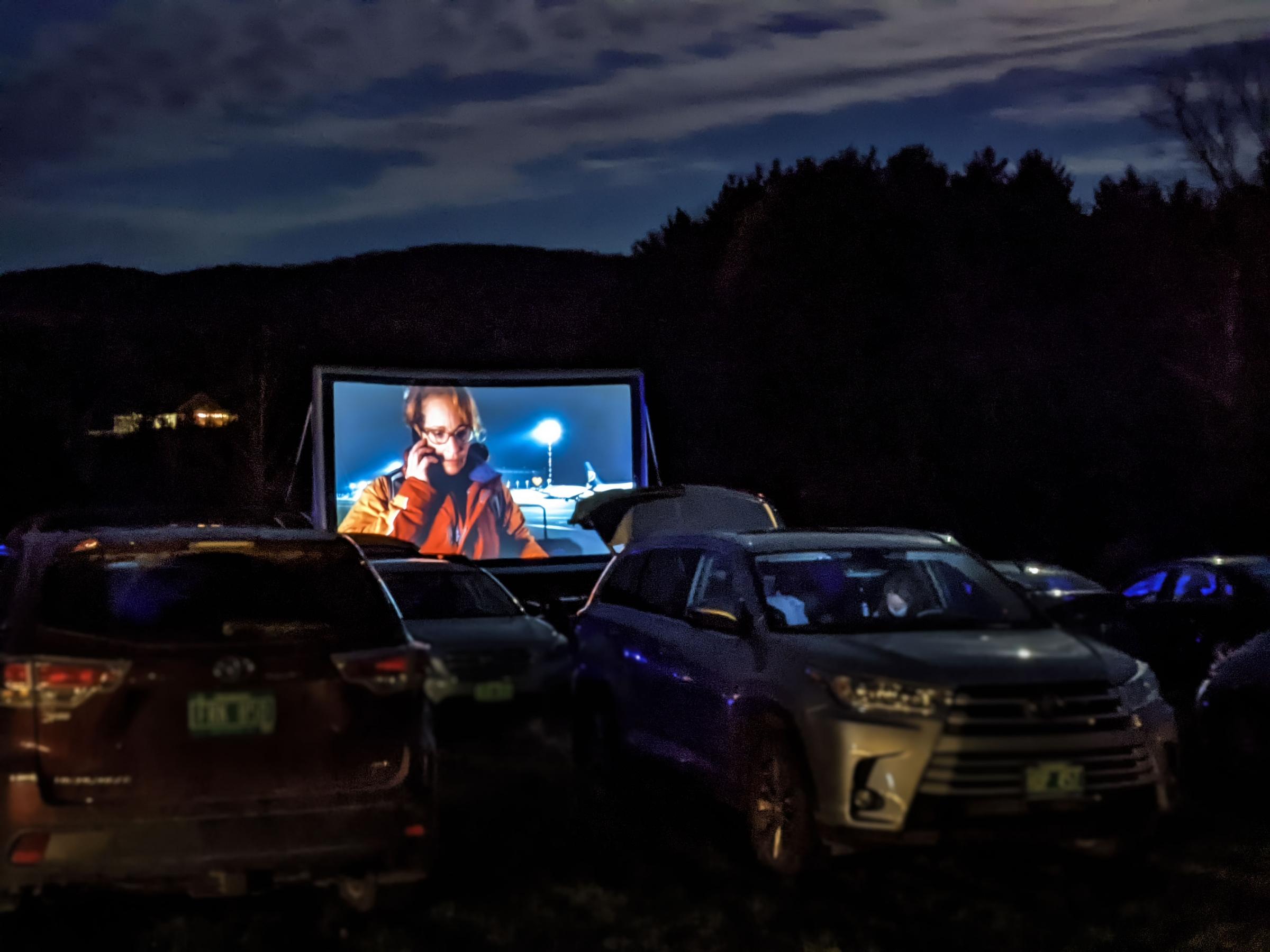 Drive in theater with cars in lot and a woman on the screen talking on the phone