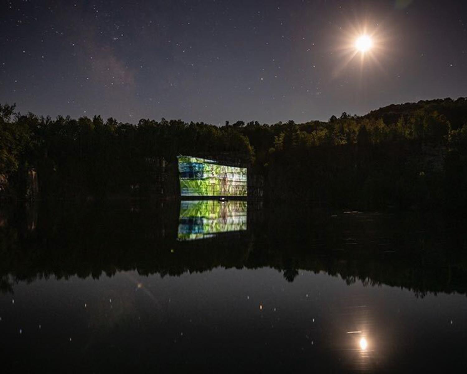 a wide shot of a quarry with a projection on the cliff wall and the moon overhead