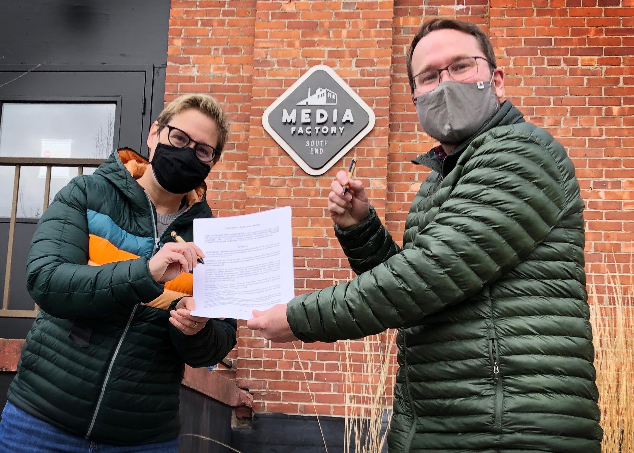 Jess and Seth sign the merger document in front of the Media Factory sign