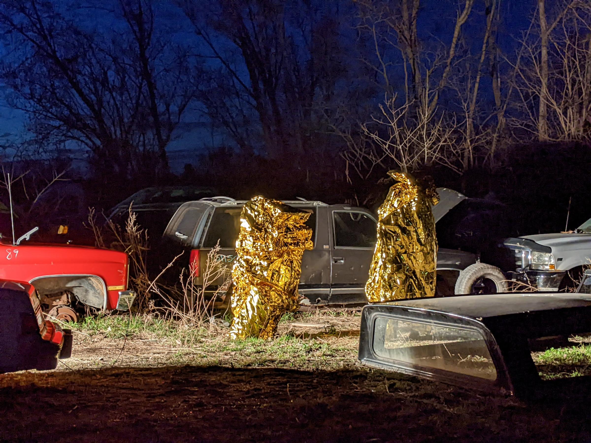 Two people wrapped in gold foil move slowly in a car junkyard