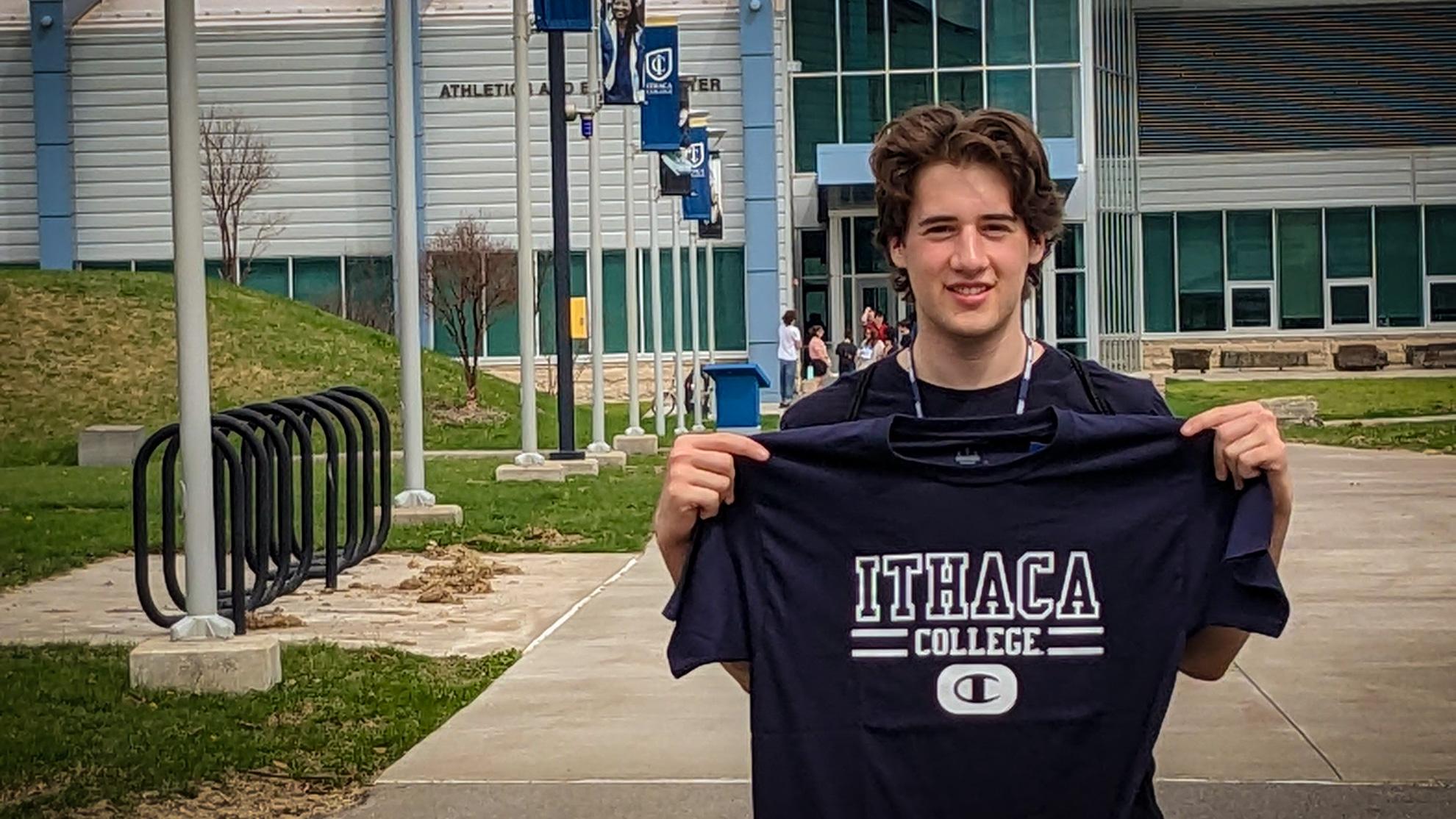 Caleb Ghia stands in front of the Ithaca College Athletic Center, holding up an Ithaca College T-shirt and smiling